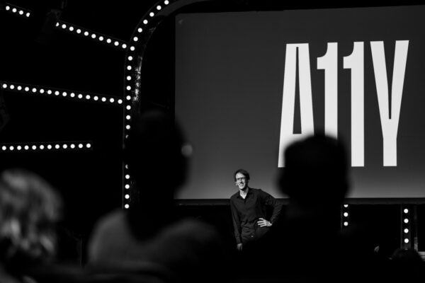 Black and white photo of Oliver Schöndorfer presenting on a stage with the term 'A11Y' displayed on a large screen, representing accessibility, with an audience in the foreground.