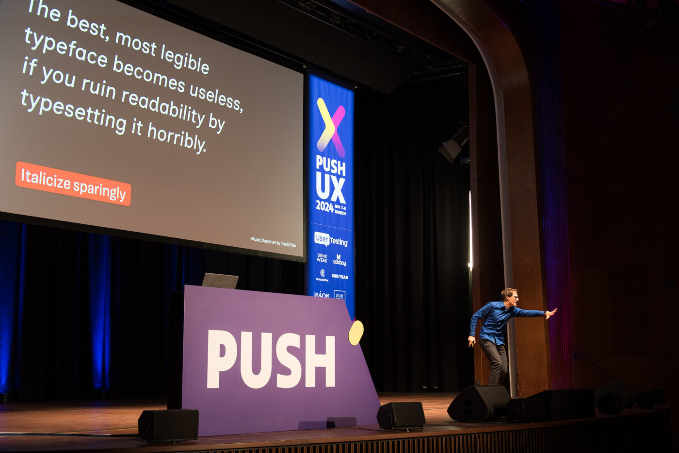 Oliver Schöndorfer presenting at PUSH UX 2024 conference with a slide about typography emphasizing readability and sparing use of italics, standing on a stage, making an engaging pose.