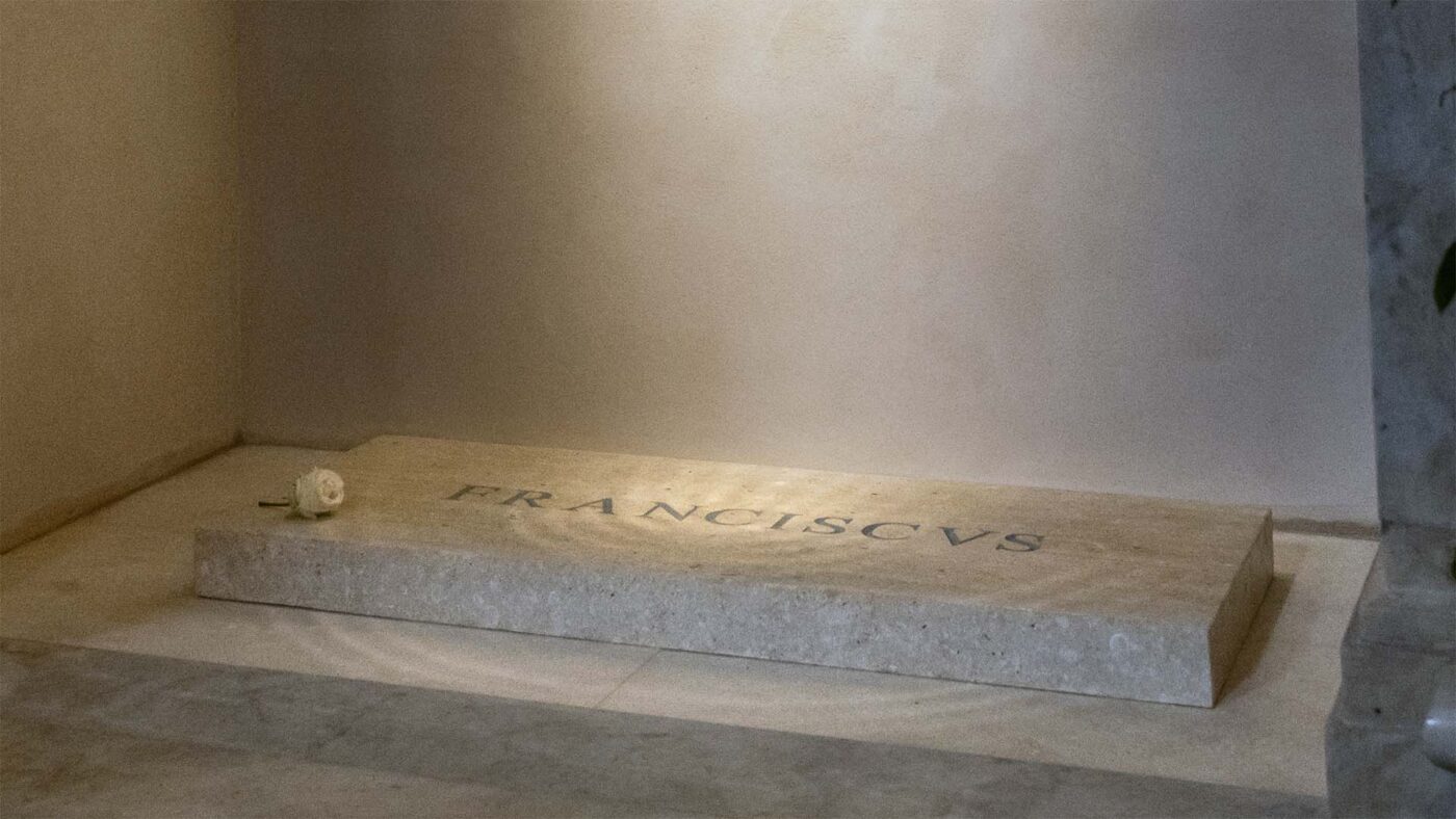 Simple stone tomb engraved with "FRANCISCVS" and a white rose placed on top, located in a quiet, minimalist chapel space.