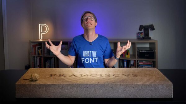 Man in a "WHAT THE FONT" shirt gestures dramatically while seated at an office desk in a workspace, with a stone slab engraved "FRANCISCVS" on the desk in front of them